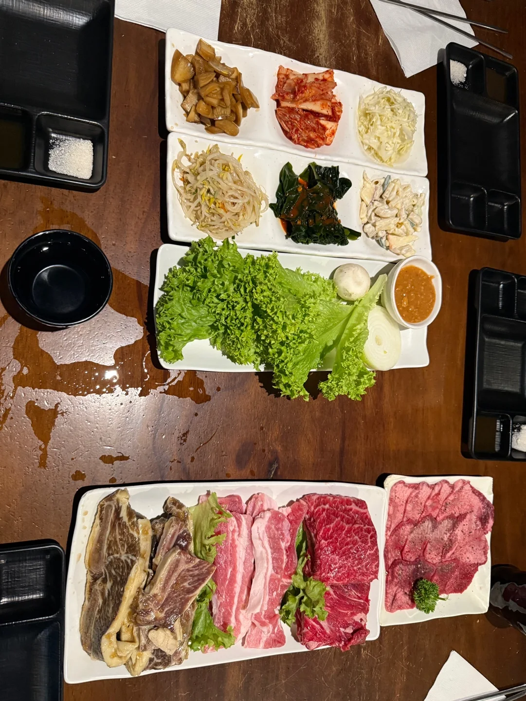 나무 테이블에 놓인 반찬 접시 두 개, 상추와 쌈장이 담긴 쌈 접시, 생고기 접시 두 개